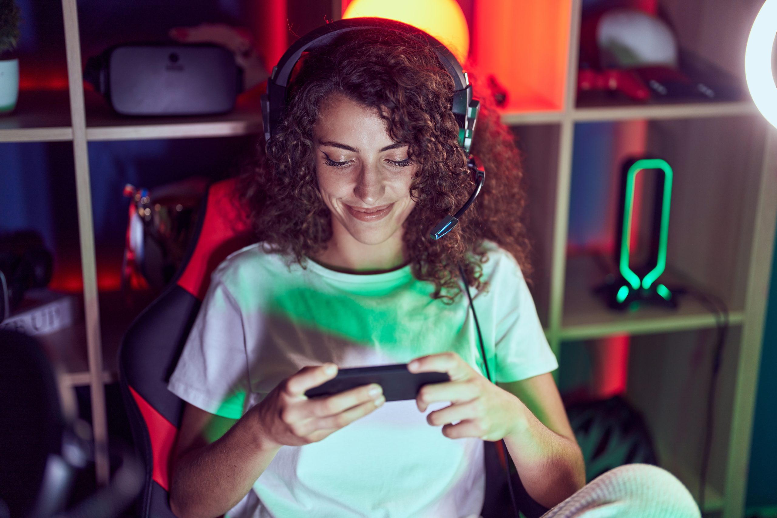 Young beautiful hispanic woman streamer playing video game using smartphone at gaming room